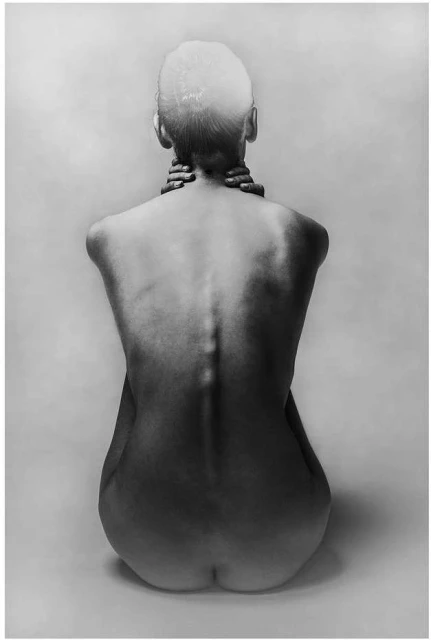 Black and white studio portrait of a bald adult woman, shot from the back, sitting and holding her neck, against a neutral backdrop.