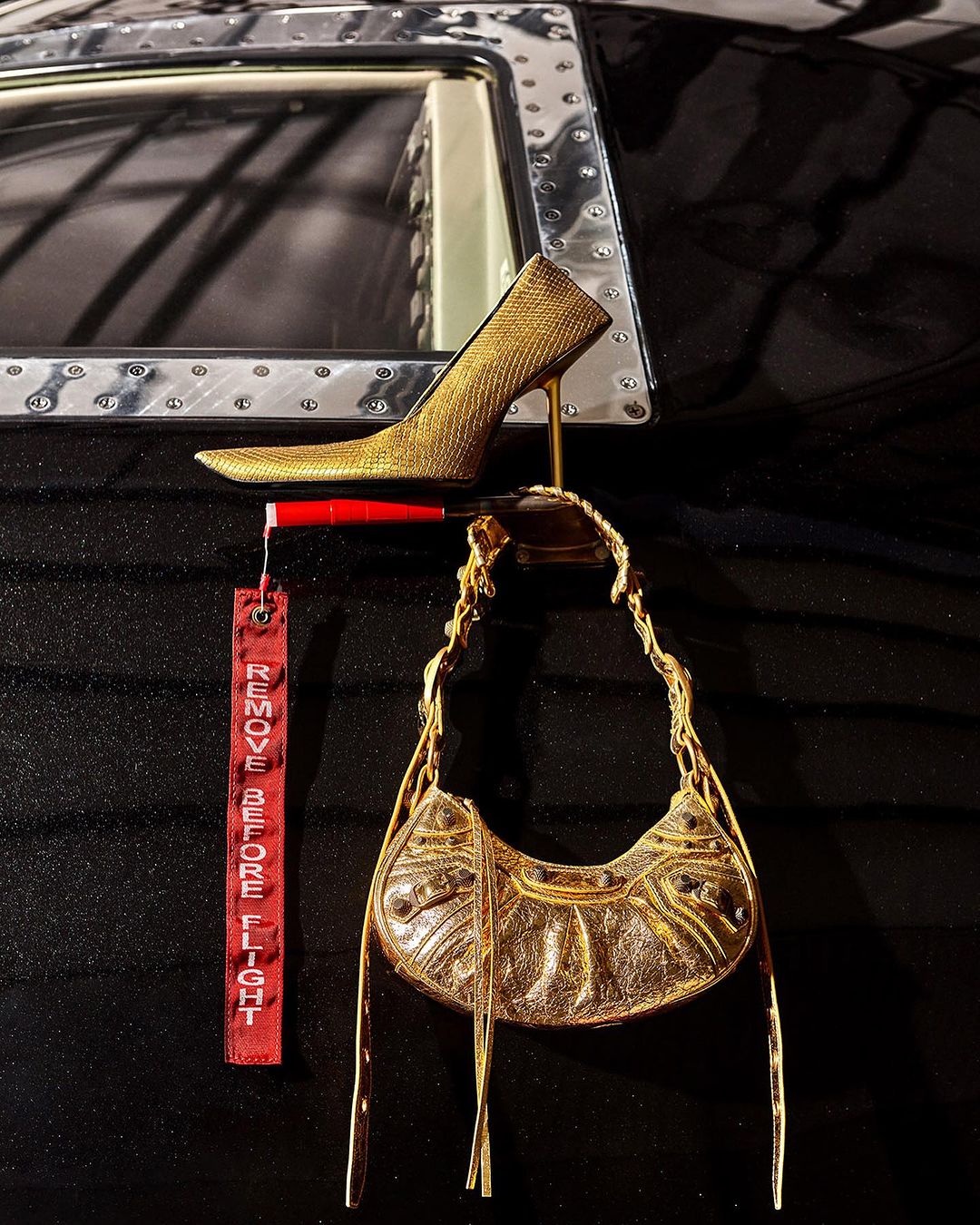Gold high heel shoe and gold purse are suspended in an airplane cabin with a red 'remove before flight' tag visible.