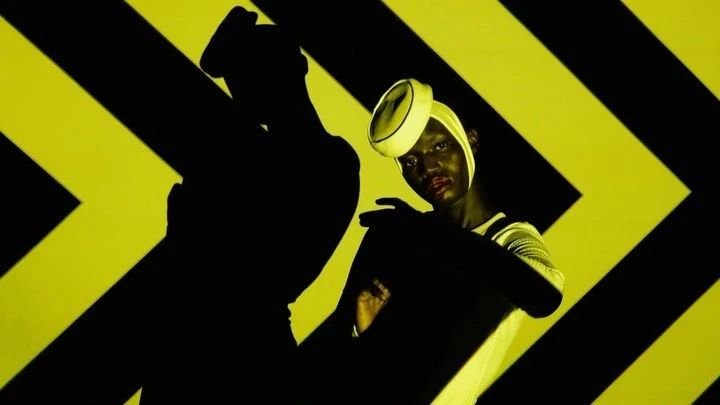 Woman with headscarf poses in front of a background of black and yellow chevron stripes, with a shadowy human figure to the left.