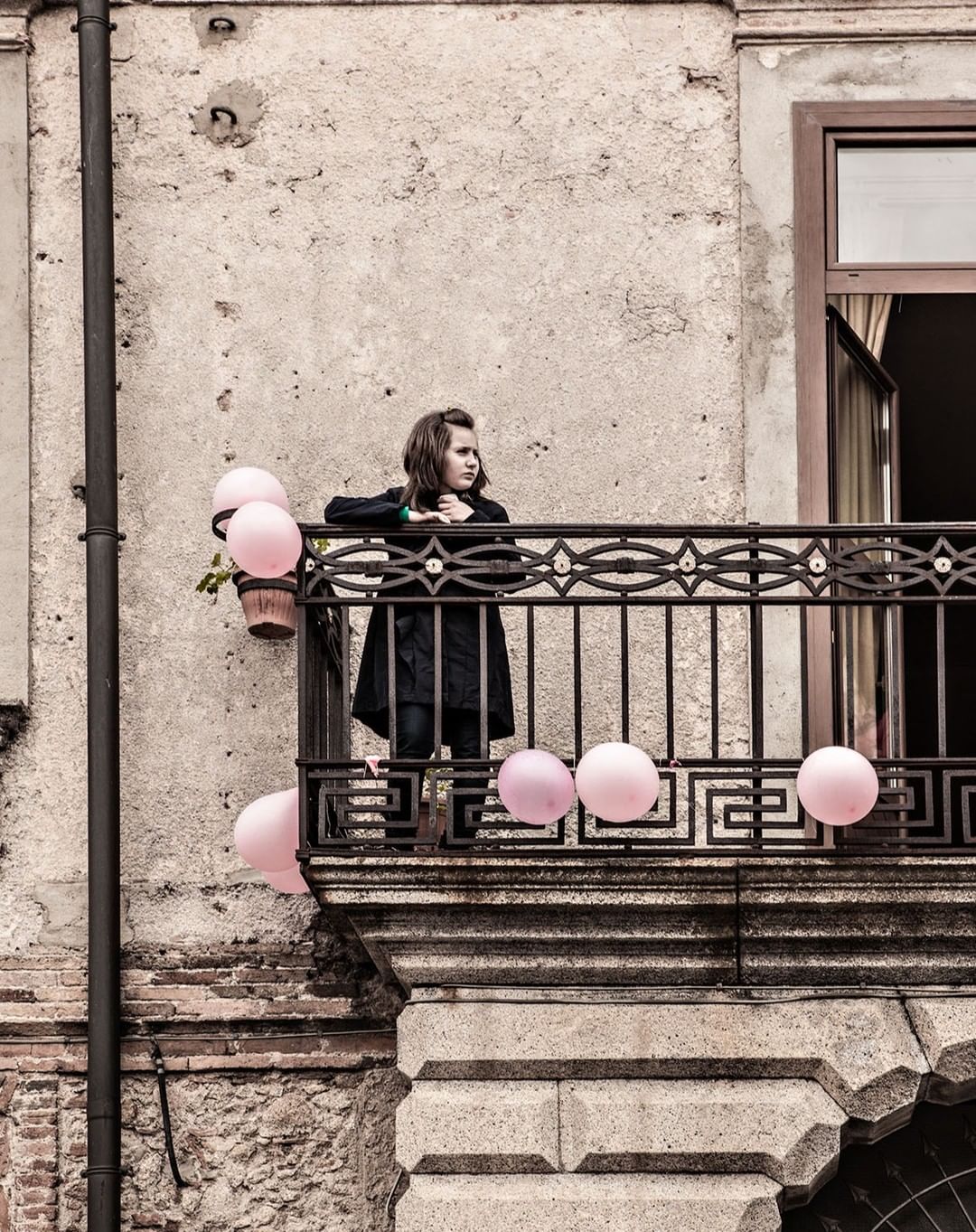 A young girl stands on a weathered balcony, surrounded by pastel pink balloons, with an aged building facade as a backdrop in daylight.