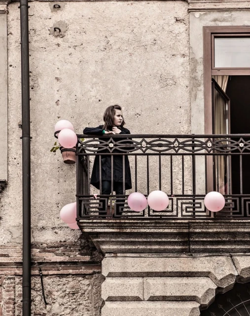 A young girl stands on a weathered balcony, surrounded by pastel pink balloons, with an aged building facade as a backdrop in daylight.