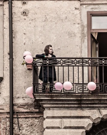 A young girl stands on a weathered balcony, surrounded by pastel pink balloons, with an aged building facade as a backdrop in daylight.