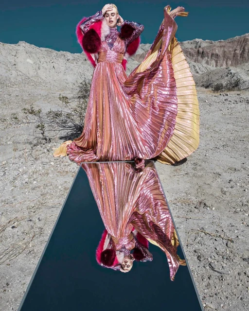 Woman in shimmering pink gown with fur bolero poses on reflective platform in arid landscape under clear sky, fashion editorial.