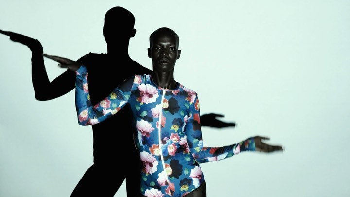 A bald African-American woman poses in a floral-patterned bodysuit with expressive hands, complemented by stark black shadows against a white backdrop.