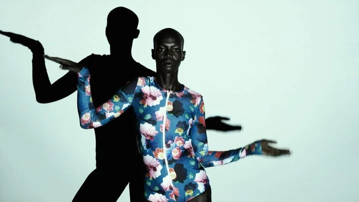 A bald African-American woman poses in a floral-patterned bodysuit with expressive hands, complemented by stark black shadows against a white backdrop.