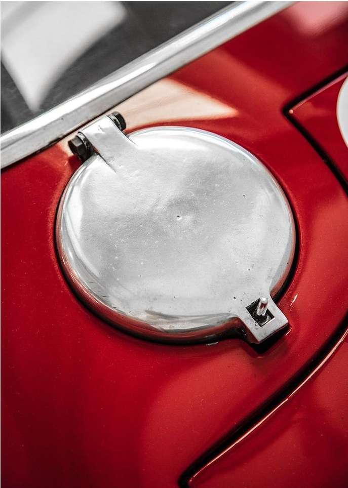 Close-up of the fuel cap of a red vintage car, with a shiny silver circular cap and visible bolts and hardware.
