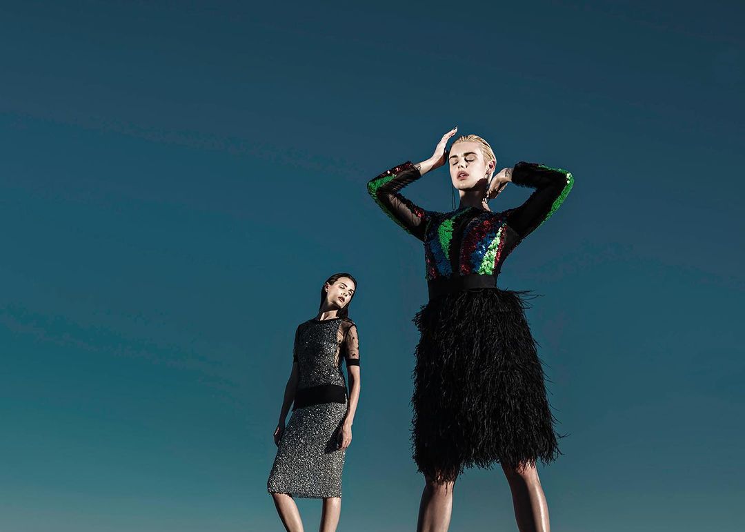 Two women in fashionable dresses against a deep blue sky. One woman is closer, wearing a sequined top and feathered skirt.
