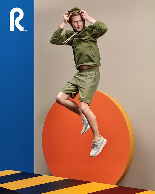 Athletic adult man in motion, mid-jump wearing a green hooded sweatshirt, matching shorts, and grey athletic shoes against a studio backdrop.