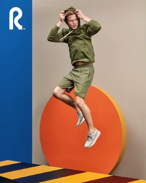 Athletic adult man in motion, mid-jump wearing a green hooded sweatshirt, matching shorts, and grey athletic shoes against a studio backdrop.