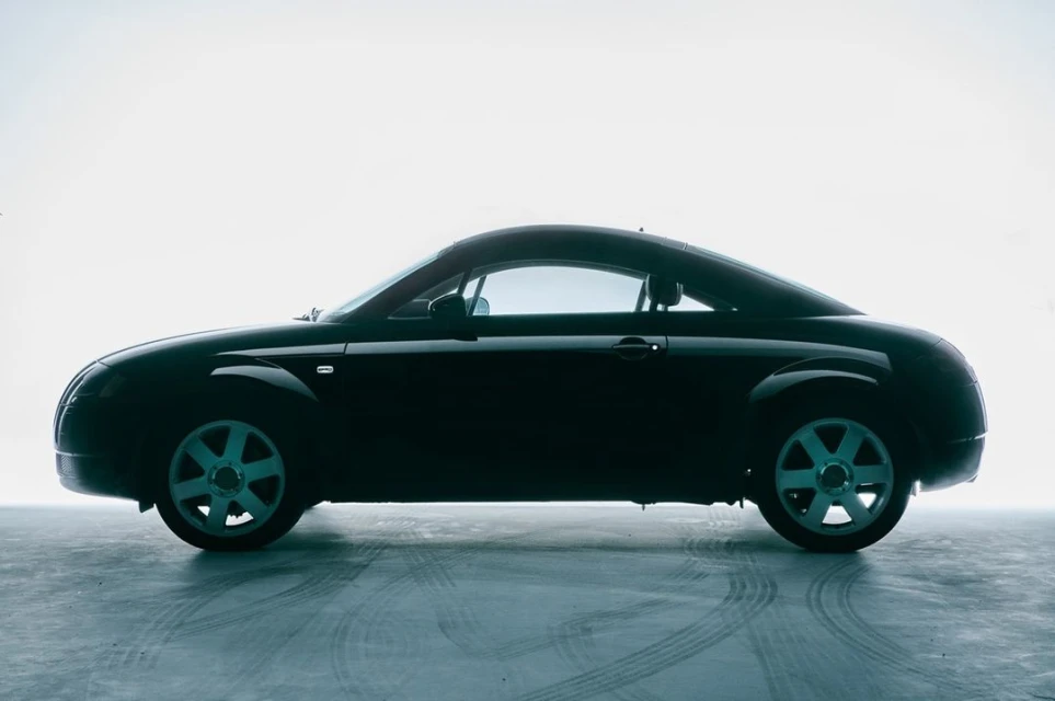 Side view of a dark grey Audi TT coupe against a bright, white background, showcasing its sporty design and low profile.