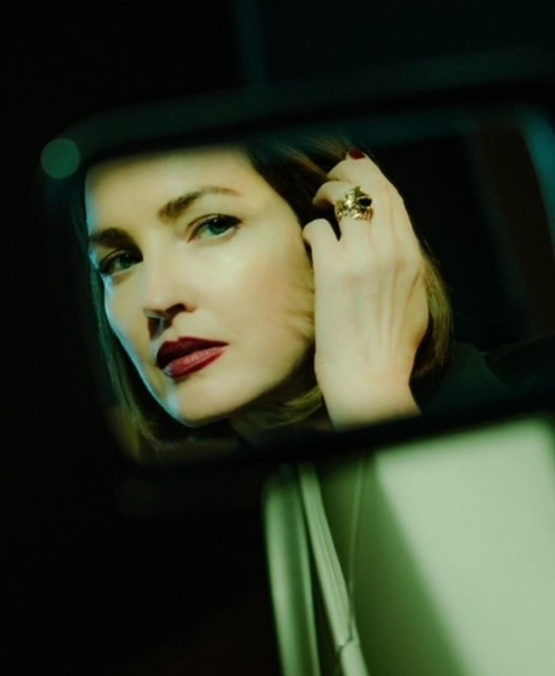 Portrait of an adult woman with blonde hair and red lipstick reflected in a car's rearview mirror at night, wearing a statement ring.