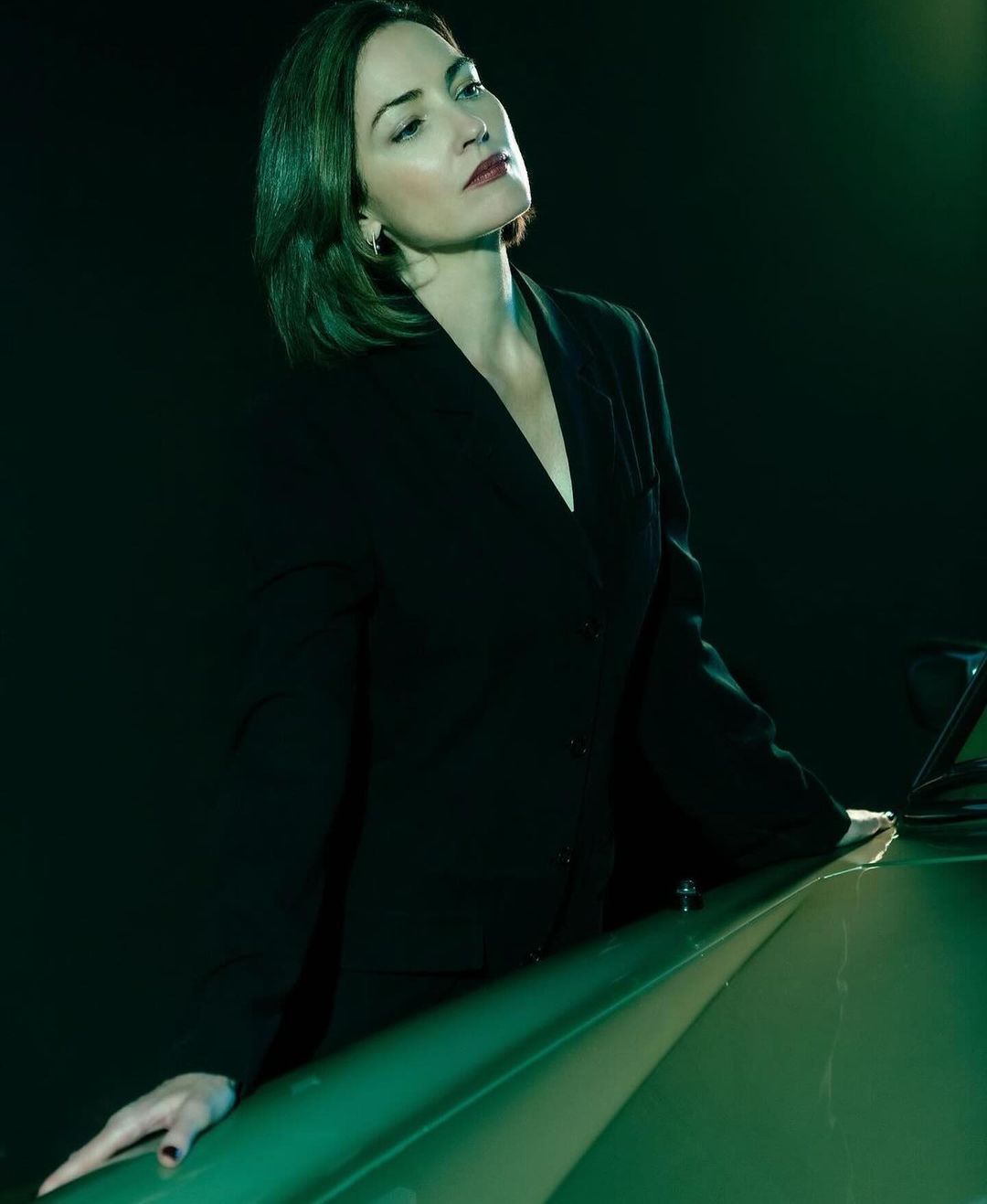 Portrait of a woman with short brown hair wearing a dark business suit jacket, posing in front of a car in low green lighting.