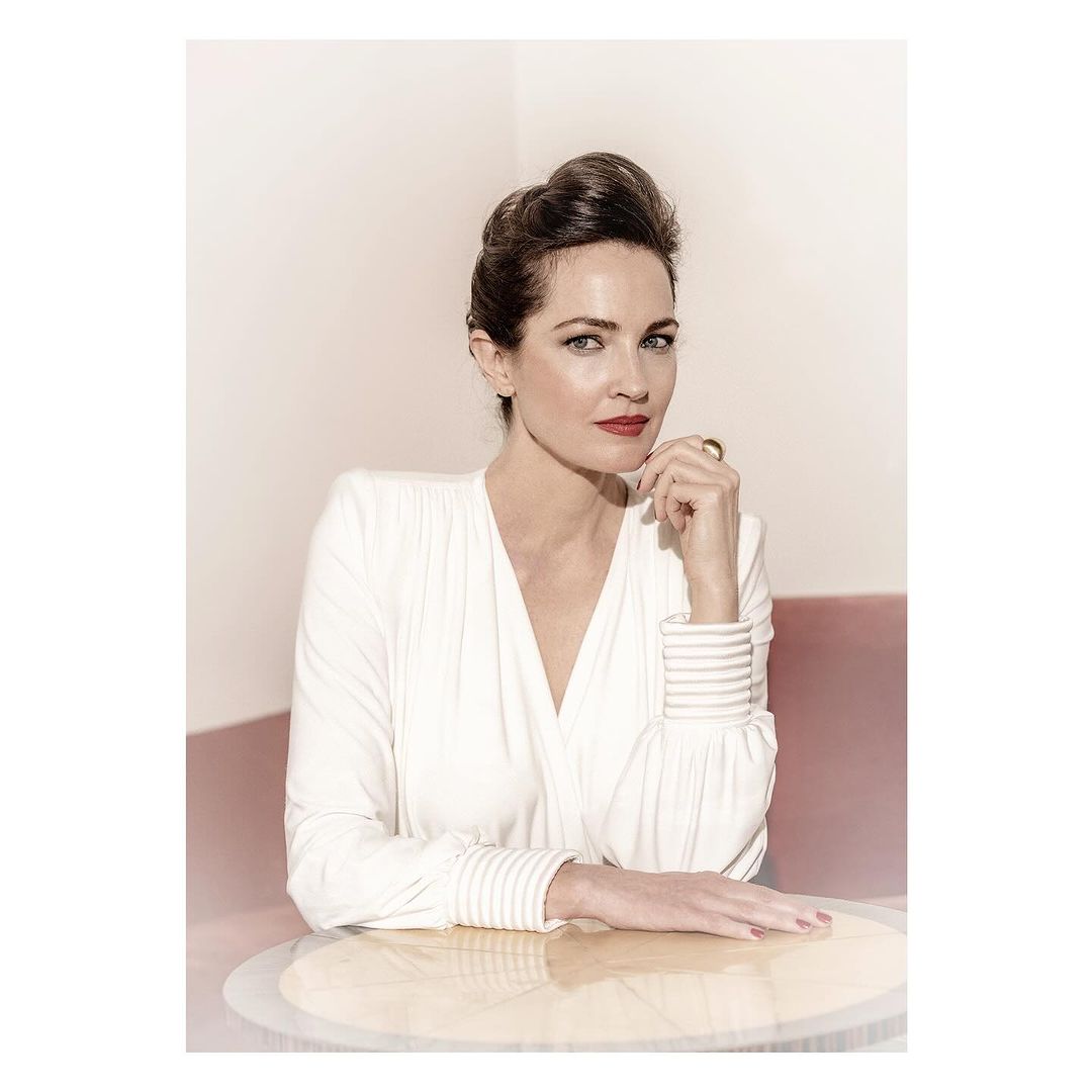 Headshot of an adult woman in a white long-sleeve top with red lipstick, posing indoors with hand near face. Light pink and white background.