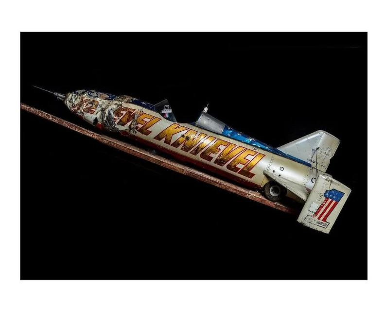 Evel Knievel toy rocket on a track against a black background. The toy rocket is white and orange with American flag decals.