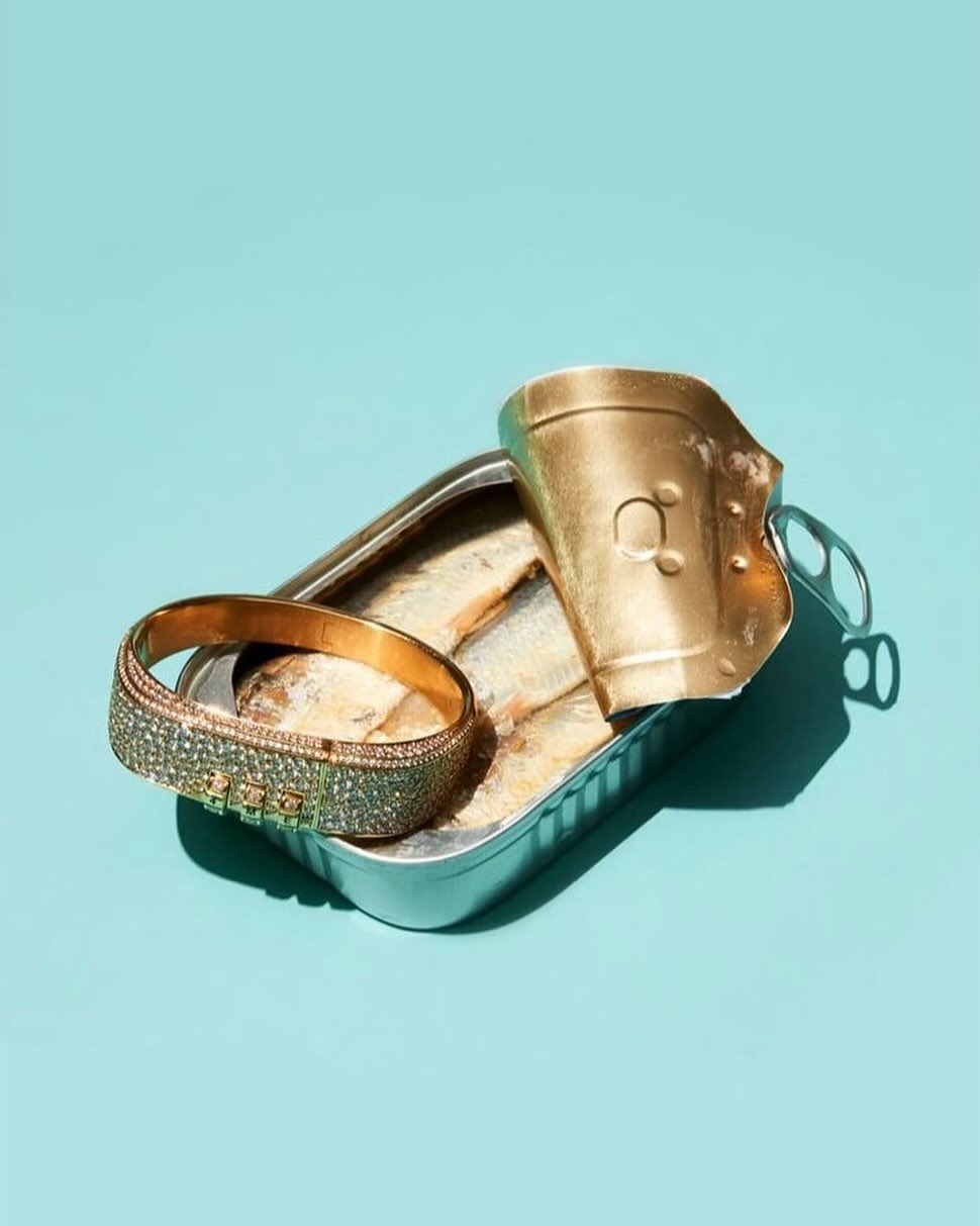 Still life of a gold bracelet placed on top of an open can of sardines against a bright blue background, illuminated with hard lighting.
