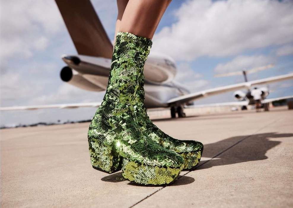 Sequin-covered green platform boots are prominently displayed against a backdrop of private jets on an airfield runway, fashion-forward.