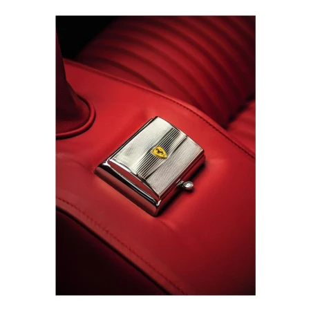Close-up shot of a silver, rectangular box with the Ferrari logo on a red leather car seat. The composition highlights luxury and automotive details.