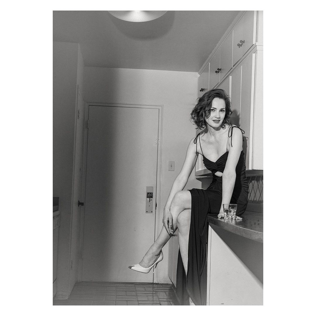 A woman sits elegantly on a kitchen counter in a black dress and heels next to a glass of liquid, creating a film-noir mood.