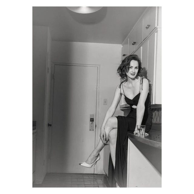 A woman sits elegantly on a kitchen counter in a black dress and heels next to a glass of liquid, creating a film-noir mood.