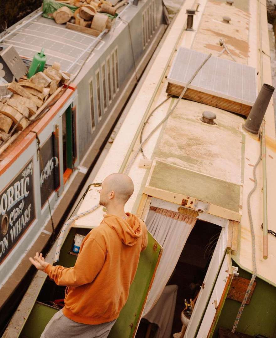 A bald man in an orange hoodie standing on a weathered boat, next to another boat loaded with logs; rustic river life.