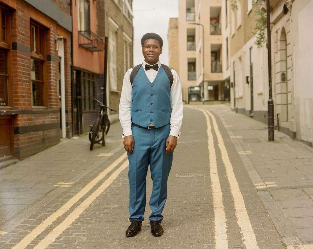Young man in teal suit and bow tie standing in the center of an urban cobblestone street lined with brick buildings and parked bicycles.