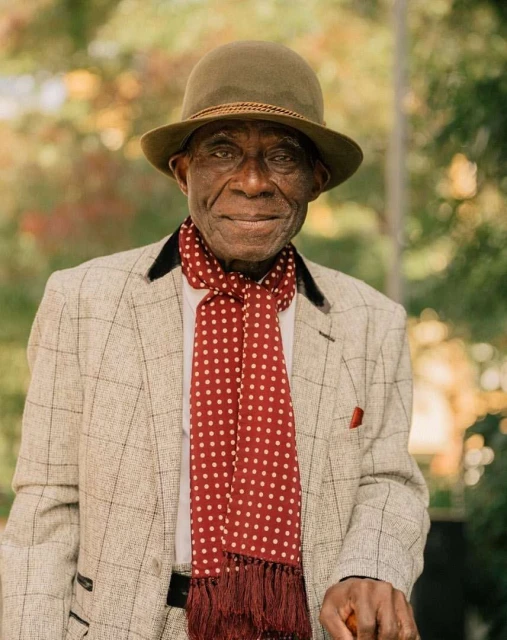Elderly African-American man in a stylish grey plaid suit, hat, and polka dot scarf poses in a natural outdoor setting. 
