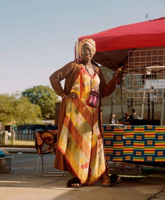 Mature woman with glasses and headscarf poses in patterned dress outdoors next to a table with patterned tablecloth.