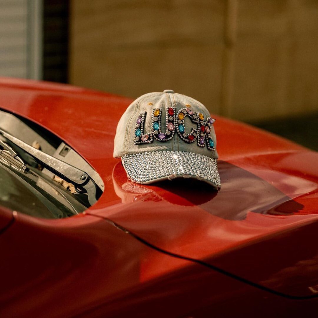 Denim baseball cap with sparkly 'LUCK' lettering resting on the red hood of a classic automobile, indoors.