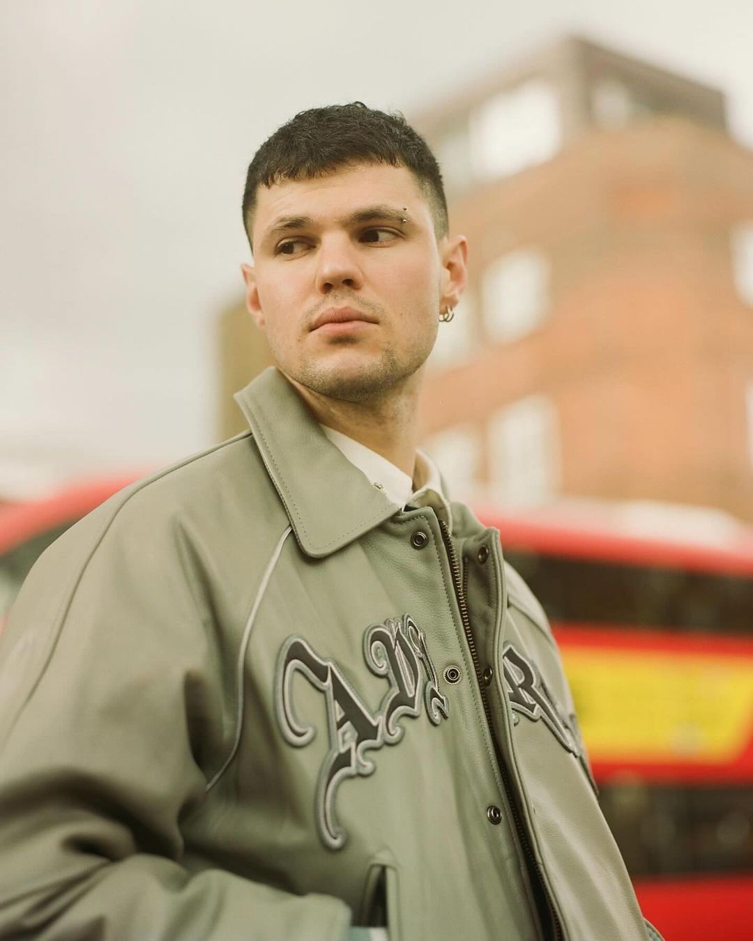 Portrait of a young adult male in a gray jacket standing outside in an urban setting with a red bus and brick building in the background.