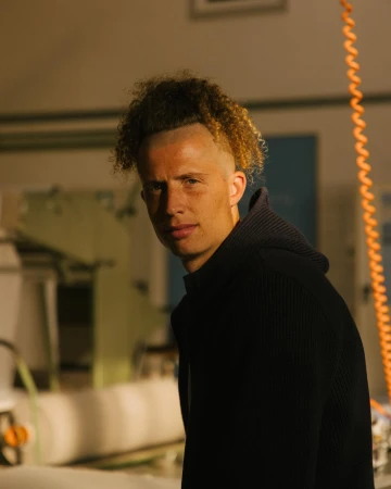 Portrait of a young adult male with curly light brown and blonde hair wearing a black hooded sweatshirt in a casual indoor setting.