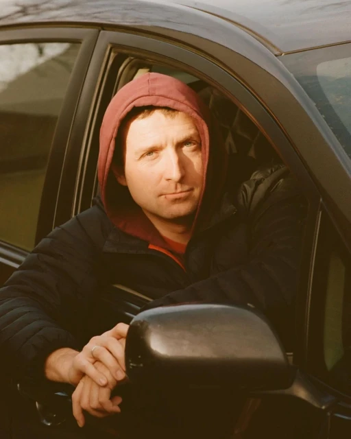 Adult male in a hooded jacket sits in a black car looking towards the camera with his arms resting on the open window.