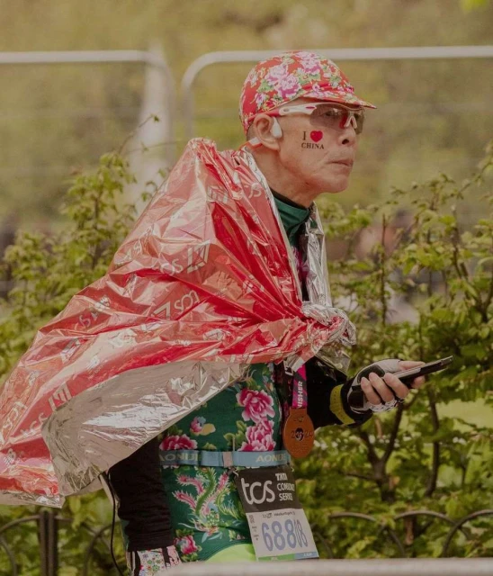 Elderly Asian marathon runner in floral pattern clothing, wearing a medal, and holding a phone outdoors near trees and metal barriers.