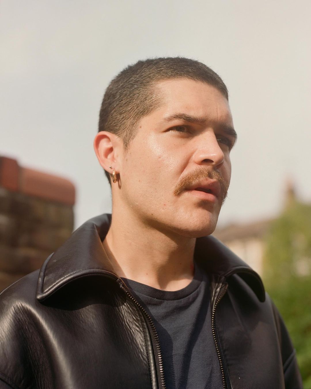 Portrait of a young adult male with a moustache, wearing a black leather jacket, against an outdoor background with trees and sky.