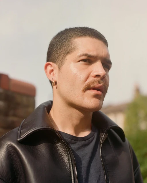 Portrait of a young adult male with a moustache, wearing a black leather jacket, against an outdoor background with trees and sky.