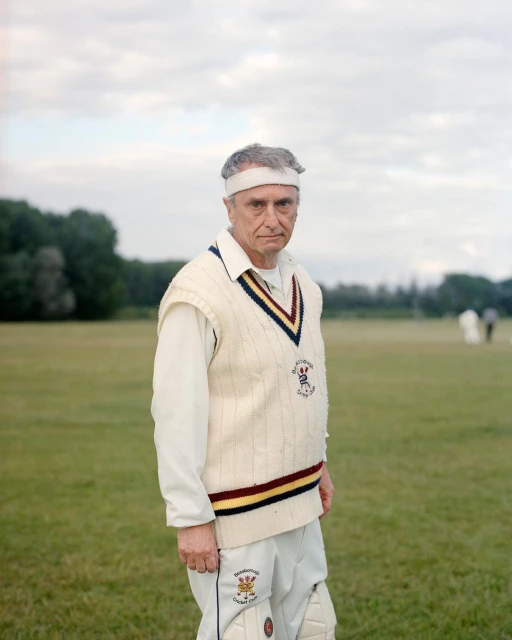 Portrait of an adult male in cricket attire, standing on a green field under a cloudy sky, wearing a cream sweater vest.