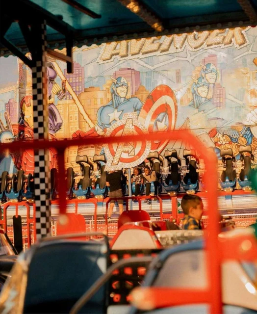 Children enjoy a spinning ride at an outdoor fair, featuring a colorful Avengers theme. The ride is in motion, under fairground lighting.