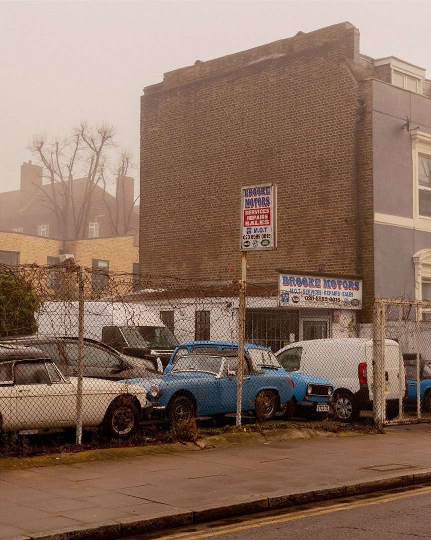 Vintage car lot featuring classic vehicles parked behind a chain-link fence, with signs for a motor repair shop against a brick building.