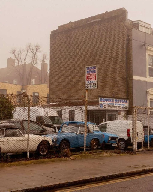 Vintage car lot featuring classic vehicles parked behind a chain-link fence, with signs for a motor repair shop against a brick building.
