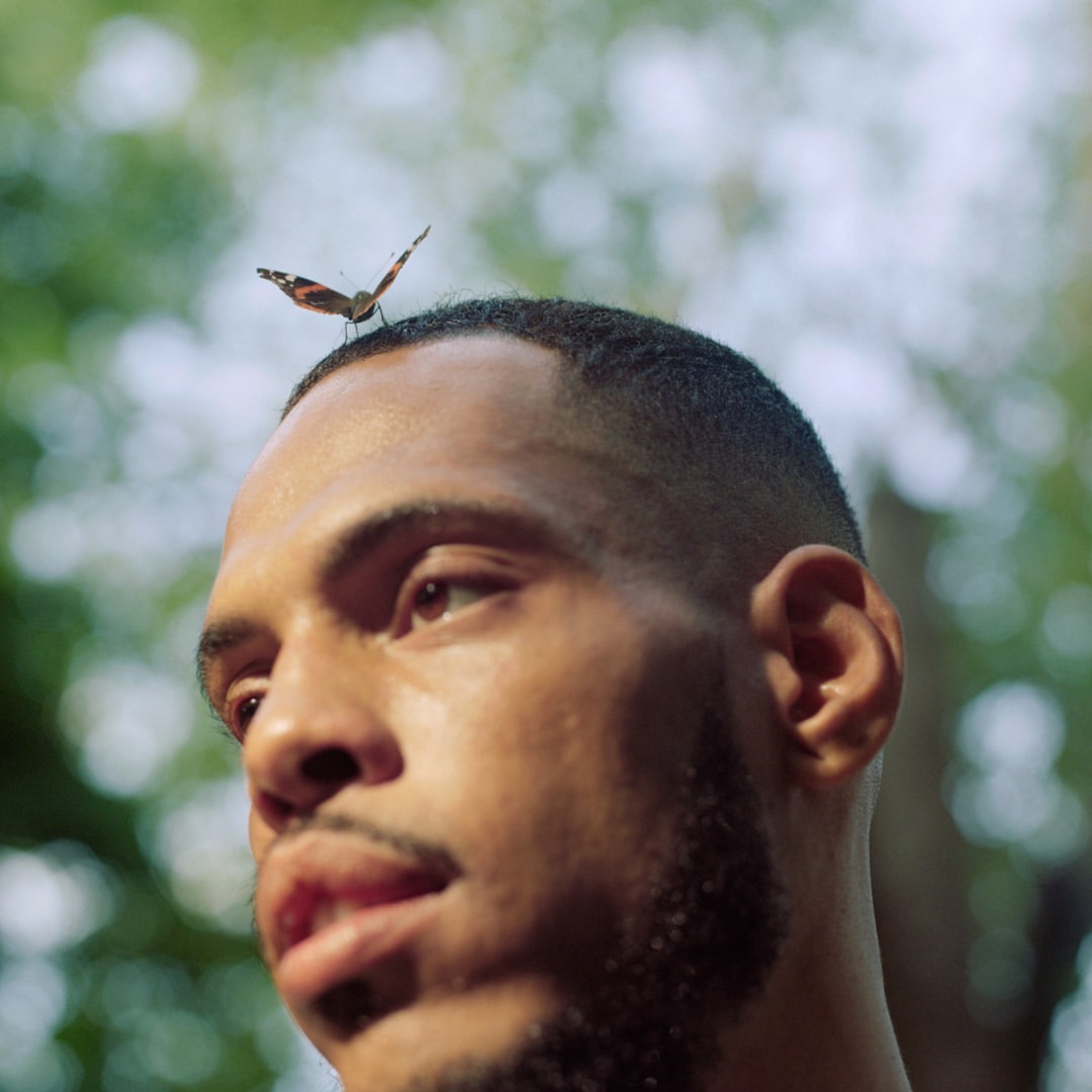 Close-up of an adult male outdoors in a blurry background, with a butterfly perched on his head. The man looks off to the side.