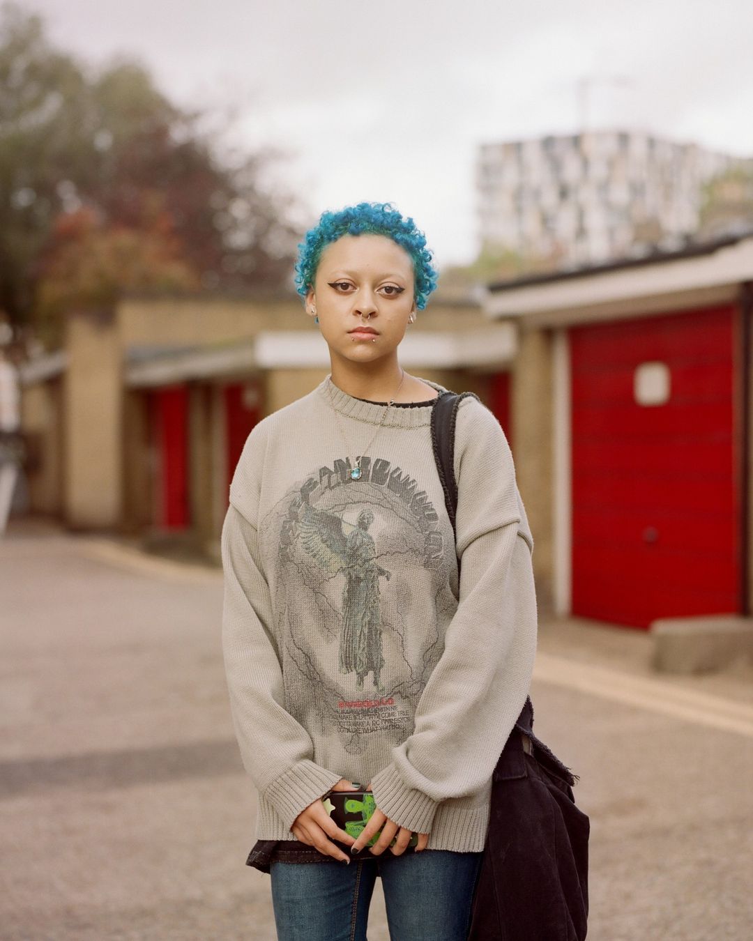 Young adult with blue hair stands outside wearing graphic sweater, jeans, and shoulder bag in an urban setting with red garage doors.