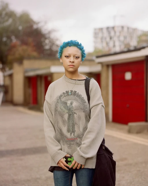 Young adult with blue hair stands outside wearing graphic sweater, jeans, and shoulder bag in an urban setting with red garage doors.