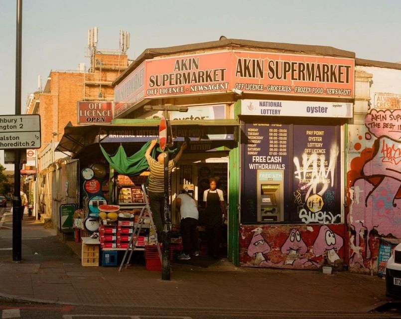 Akin Supermarket in urban area shows a man on a ladder adjusting a covering, with a street scene and brick buildings in the background.
