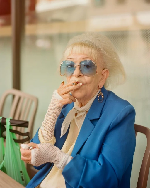 Stylish elderly woman in blue sunglasses and a blue blazer smoking a cigarette while seated at a table outdoors during the daytime.