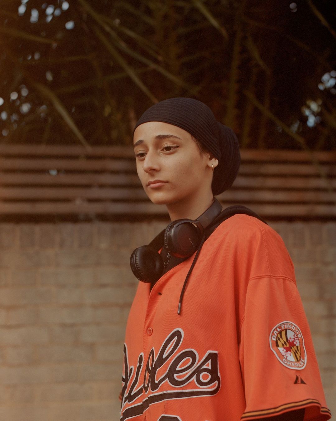 Teen girl in Orioles jersey and black beanie, wearing headphones outdoors against wall and foliage, in medium shot with natural lighting.