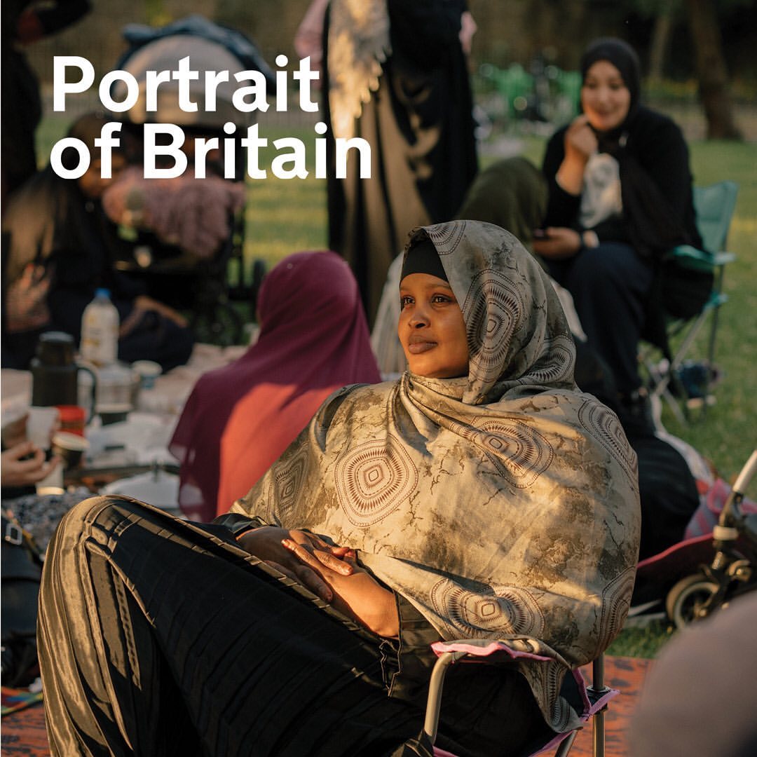 Portrait of a woman wearing a hijab, seated outdoors in a chair, surrounded by other women and greenery in soft, diffused light.