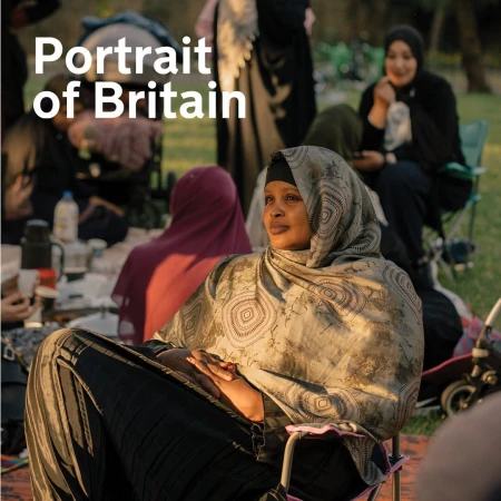 Portrait of a woman wearing a hijab, seated outdoors in a chair, surrounded by other women and greenery in soft, diffused light.