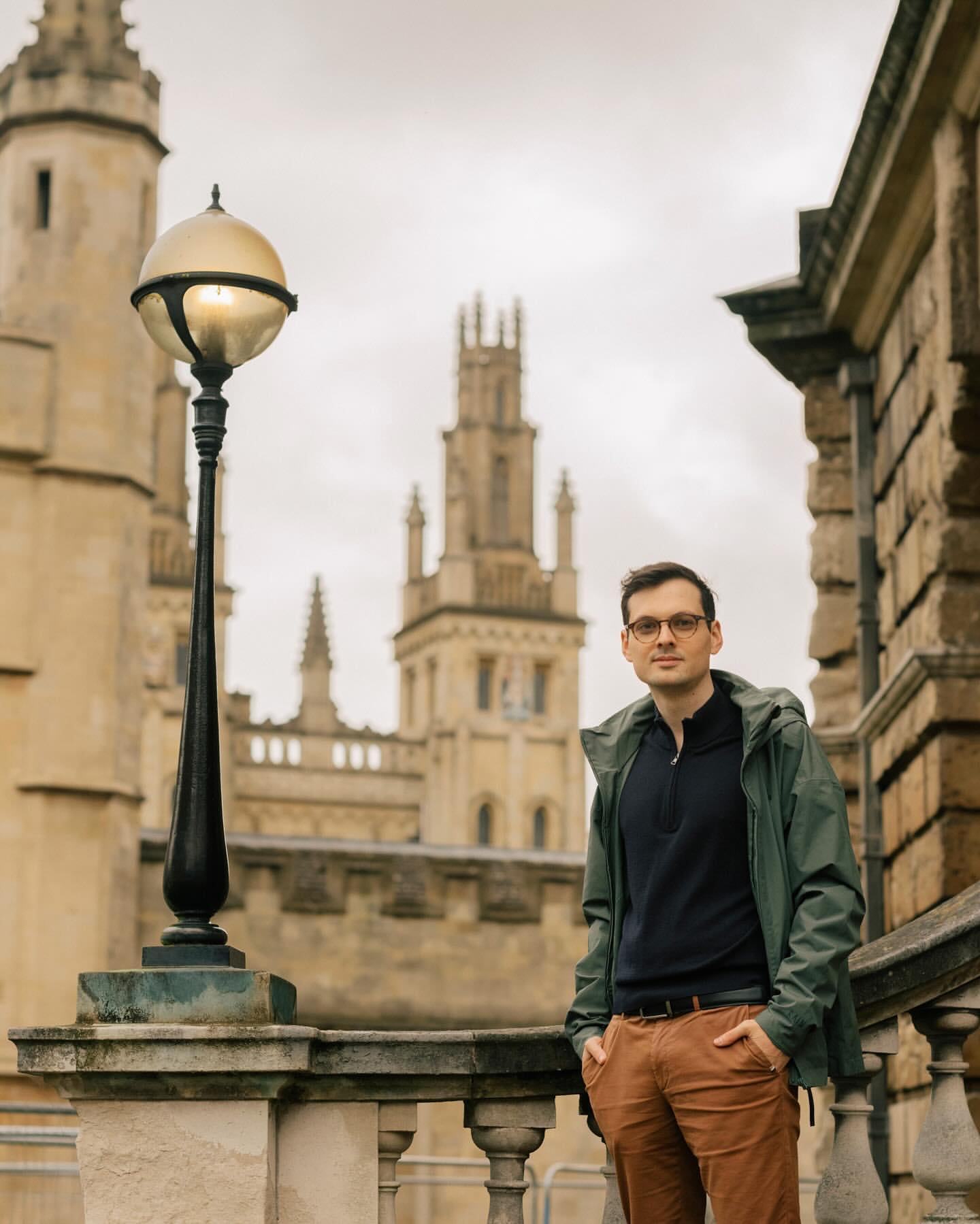 Adult male poses on stone terrace in green jacket, glasses, khaki pants, framed by vintage street light and cathedral-like buildings in background.