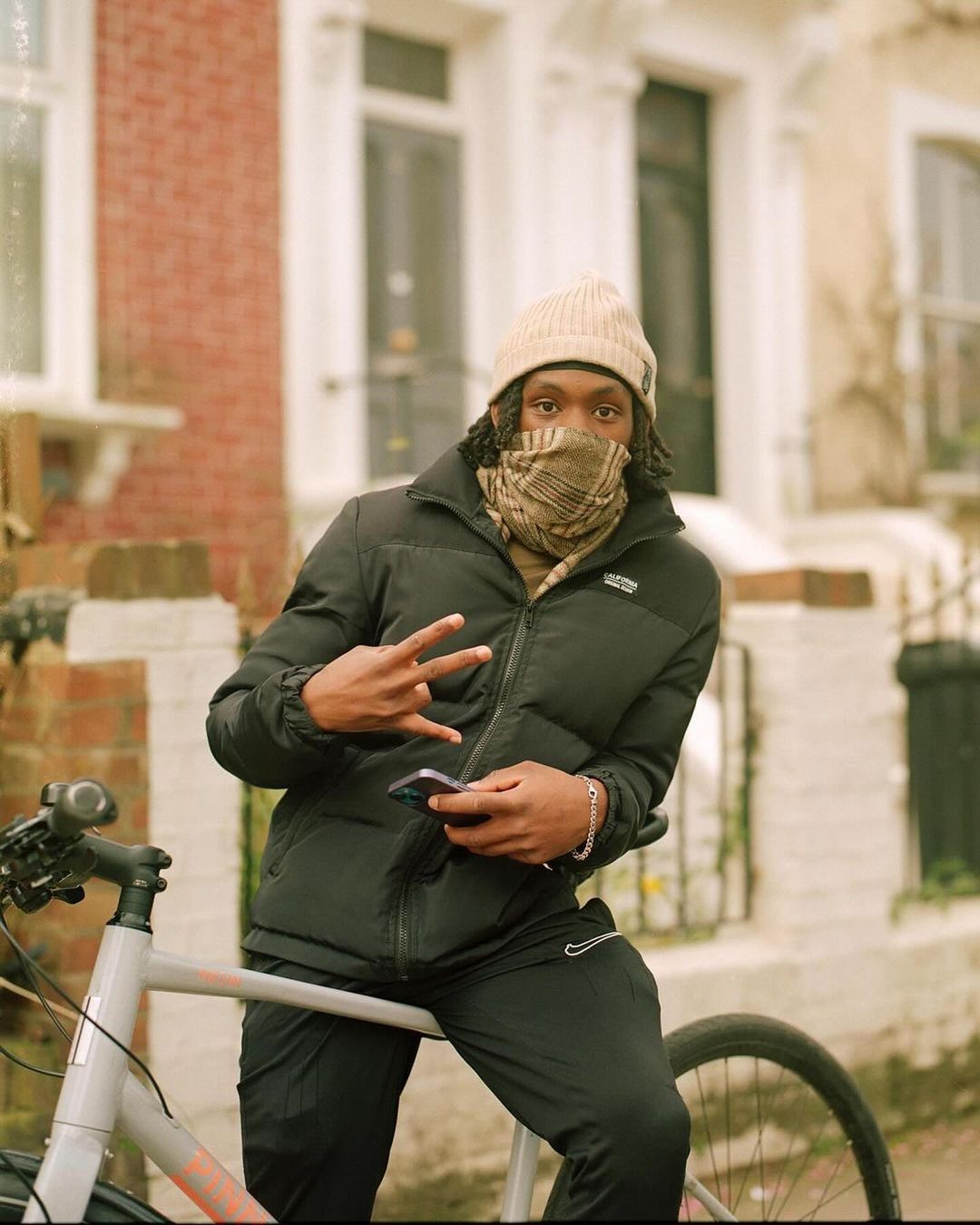 Young adult poses with bike in urban setting, wearing beanie, scarf covering face, and puffer jacket; gesturing the peace sign.
