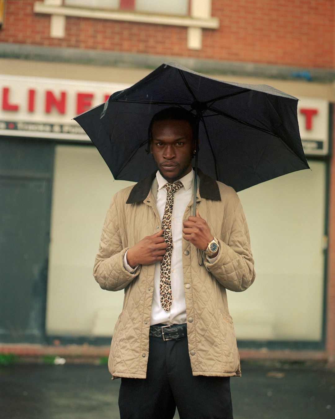 A young black man standing outside in the rain under an umbrella, wearing a beige jacket, white shirt, and patterned tie.