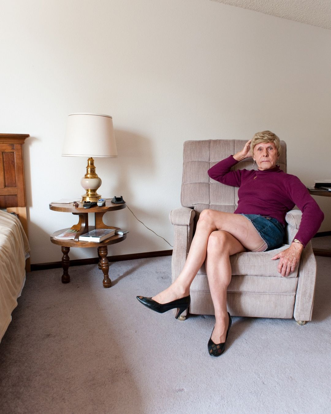 Elderly woman with short blonde hair relaxes in armchair wearing a purple sweater, blue skirt, and black high heels in a room setting.